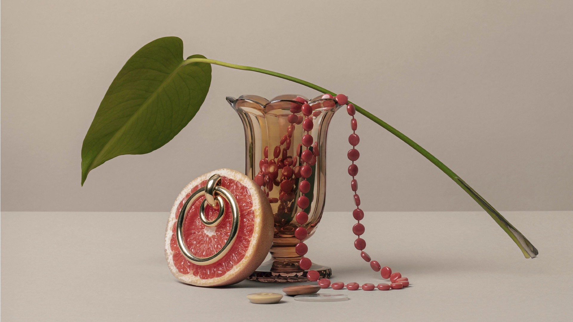 Odd objects put together. A vase with a red pearl necklace in it, a grapefruit made into a key ring and a monstera leaf - NyStudio