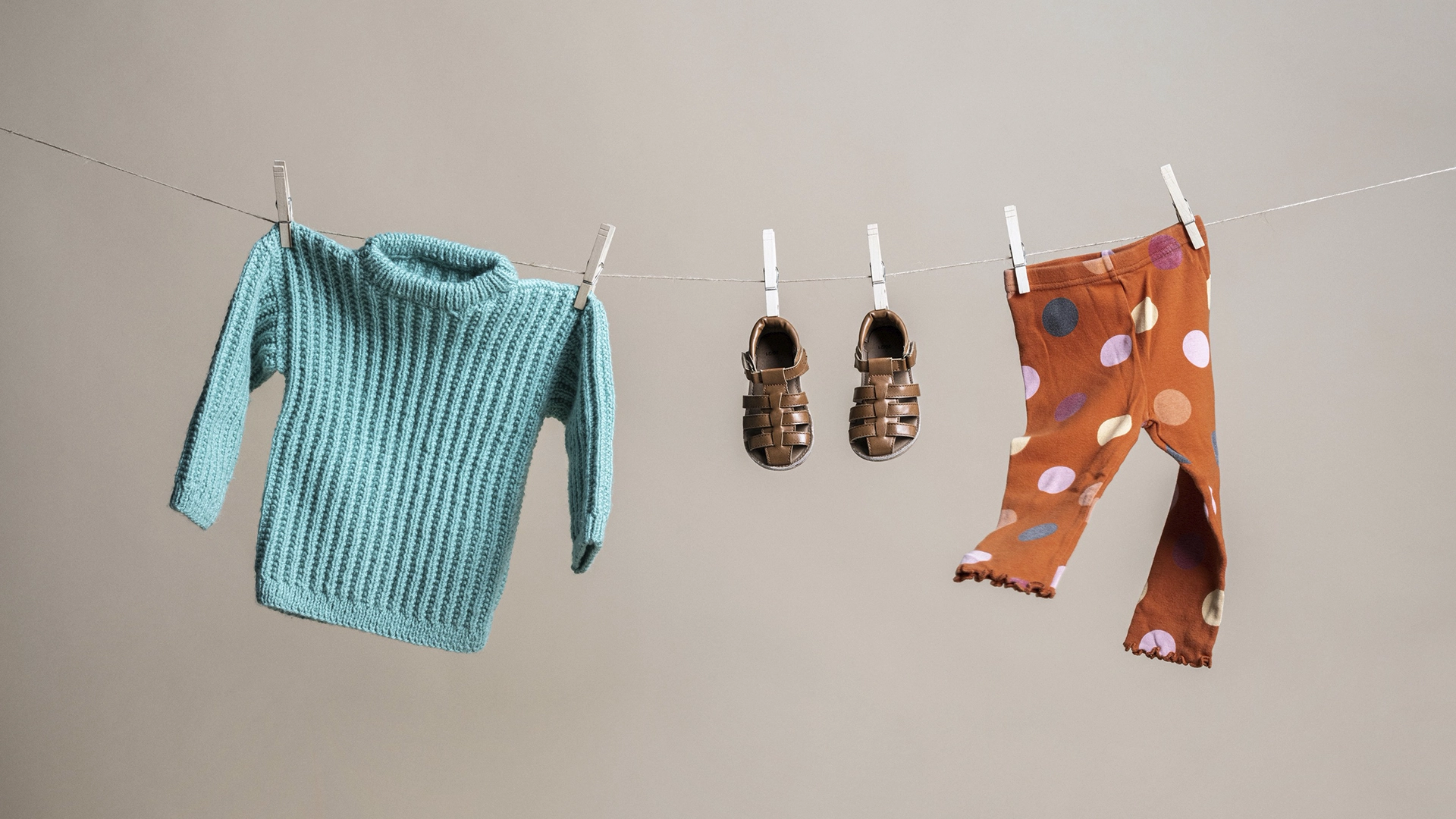 Three children's clothes a blue knitted cardigan, shoes and a pair of leggings on a clothesline attached with clothespins.